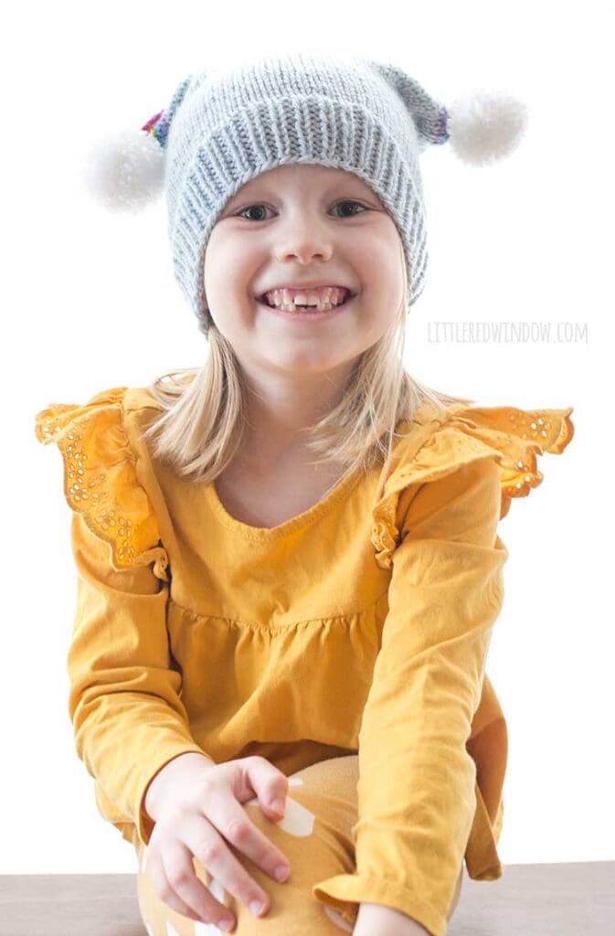 smiling girl in yellow shirt wearing blue knit hat with two white pom poms on top
