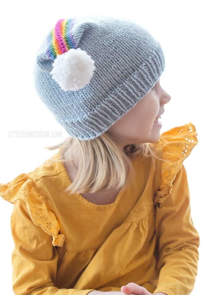 girl in yellow shirt wearing blue knit hat with rainbow on top and white pom pom on the corner looking off to the right
