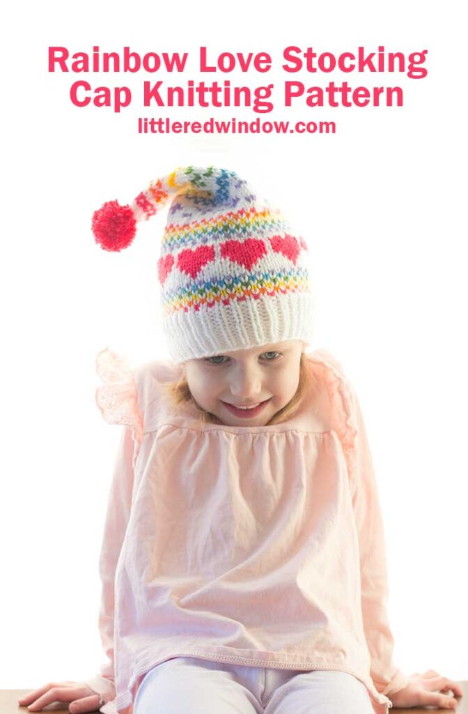 girl with shrugged shoulders wearing a white knit stocking cap with pink hearts and rainbow fair isle design