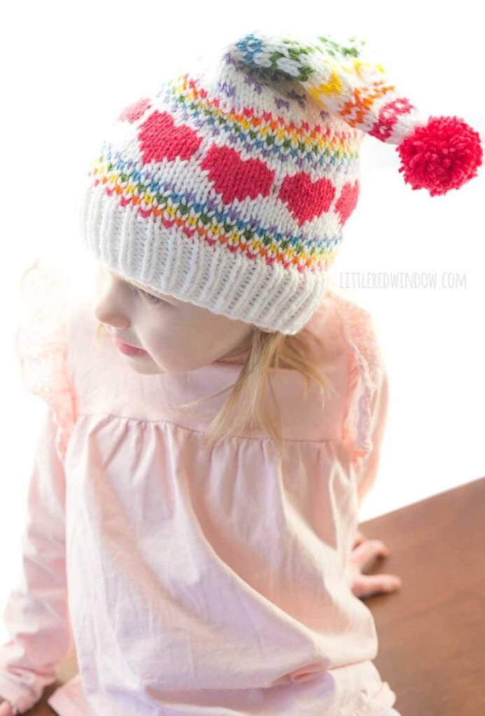 top view of girl in pink shirt wearing white and rainbow colored rainbow love stocking cap with hot pink pom pom