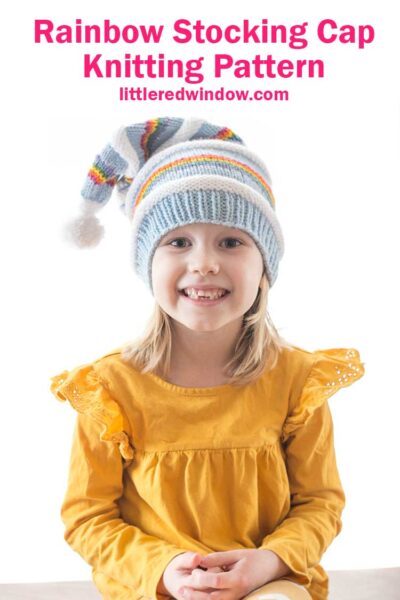 smiling girl in yellow shirt wearing a knit stocking cap with stripes of blue white and rainbow