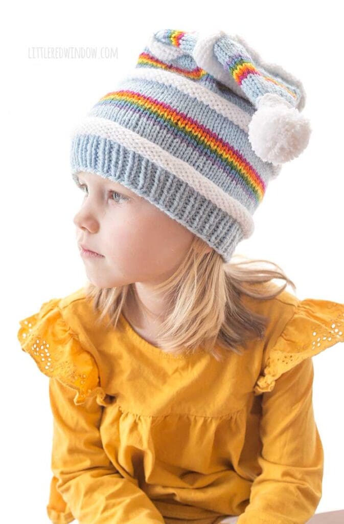girl in yellow shirt wearing knit stocking cap with rainbow blue and white stripes looking off to the left