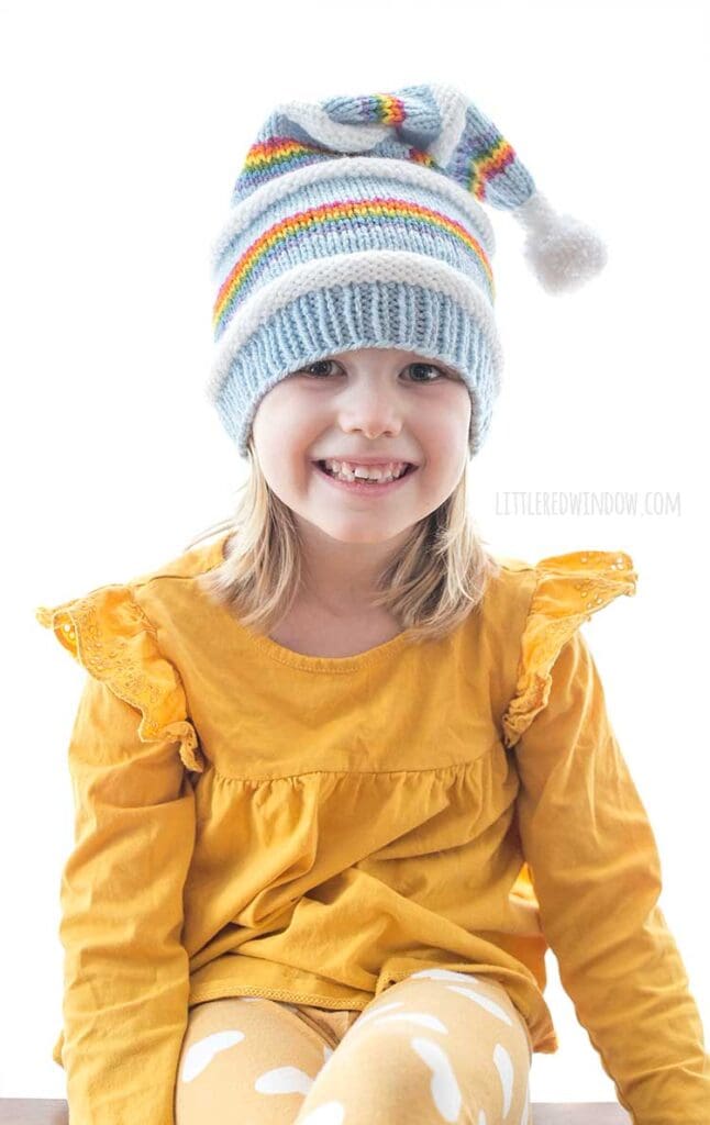 smiling girl in rainbow stocking cap with white pom pom flopped over to the right