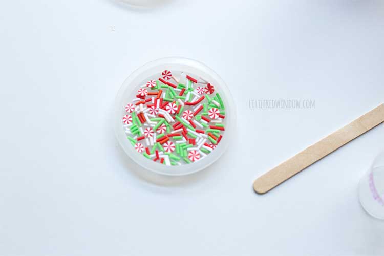 one circular flat ornament mold with red and green faux sprinkles on the bottom next to one popsicle stick on a white table