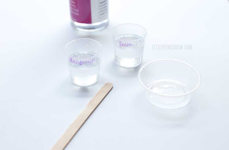 closeup of an empty container a popsicle stick and two small cups filled halfway with clear liquids labeled resin and hardener