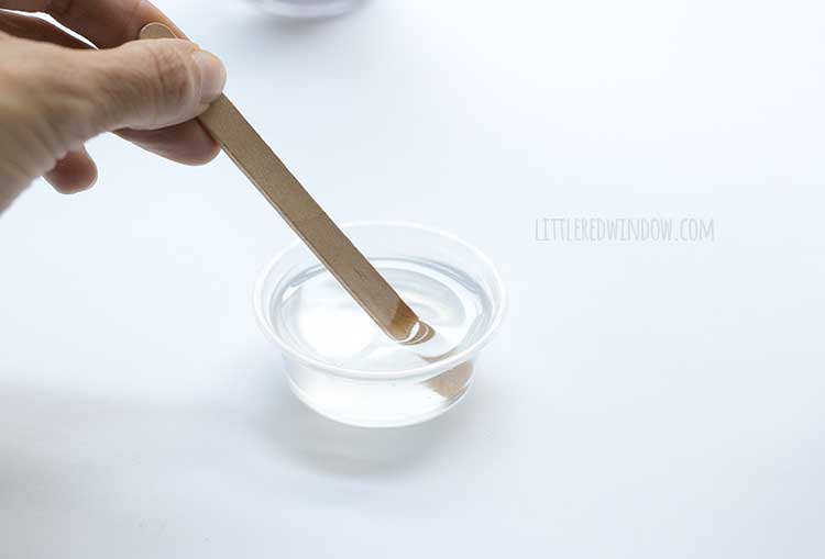 hand stirring a small container of clear liquid with a popsicle stick on a white background