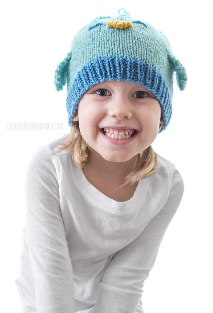 smiling girl leaning toward the camera and wearing a blue knit hat that looks like a bluebird