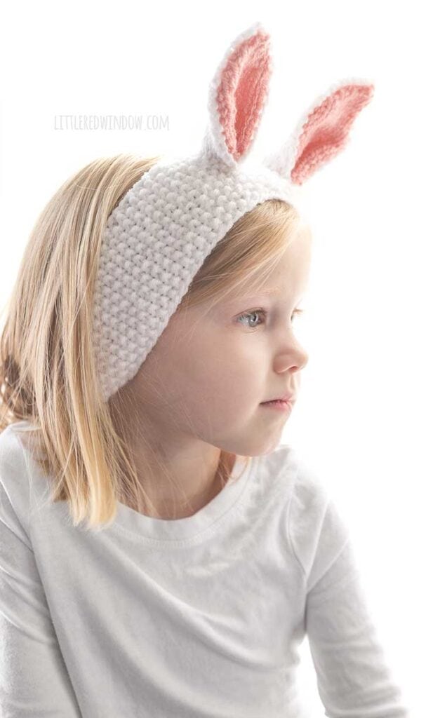closeup of little blond girl in white shirt wearing seed stitch knit white headband with white and pink bunny ears on top looking off to her right