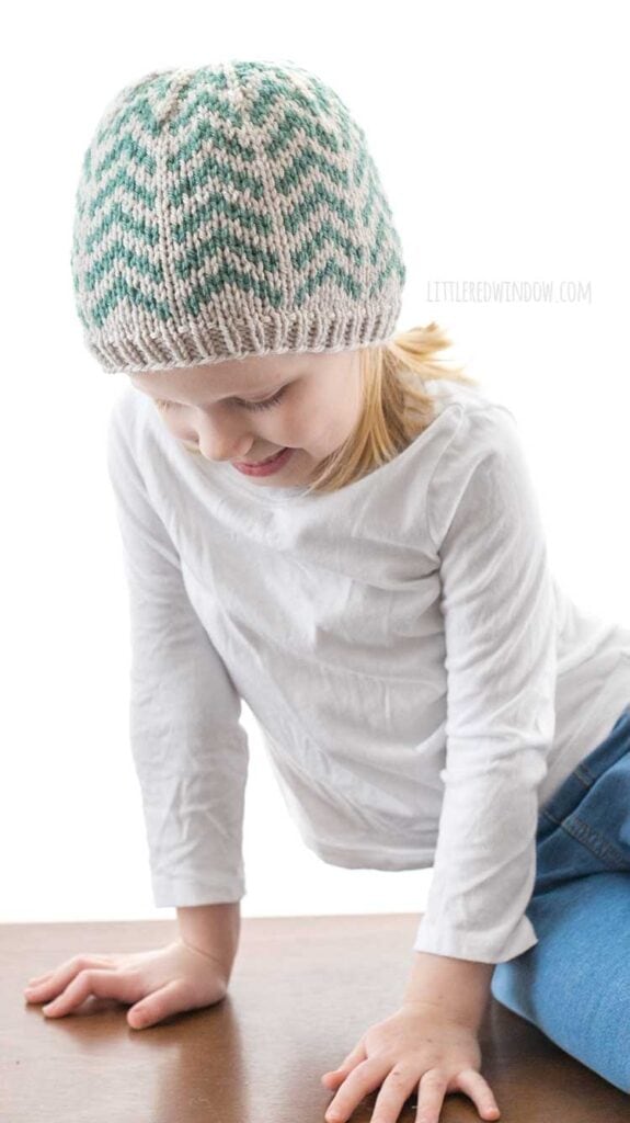 girl in white shirt leaning down and wearing tan knit hat with teal arrow pattern on it