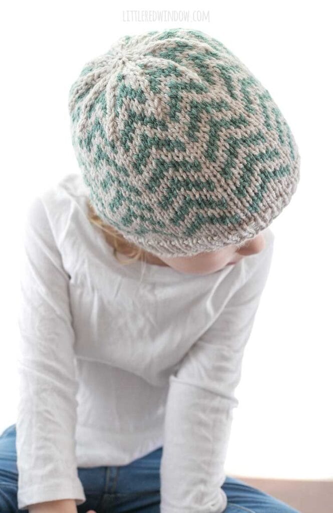 girl looking down over her shoulder and wearing tan knit hat with teal arrow shapes going up to the top