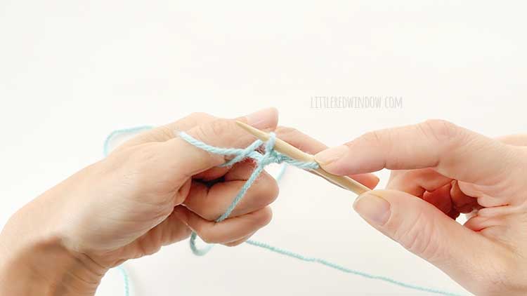 Two hands in front of a white background holding one knitting needle and light blue yarn showing german twisted cast on step 10