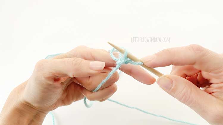 Two hands in front of a white background holding one knitting needle and light blue yarn showing german twisted cast on step 11