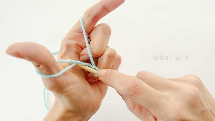 Two hands in front of a white background holding one knitting needle and light blue yarn showing german twisted cast on step 2