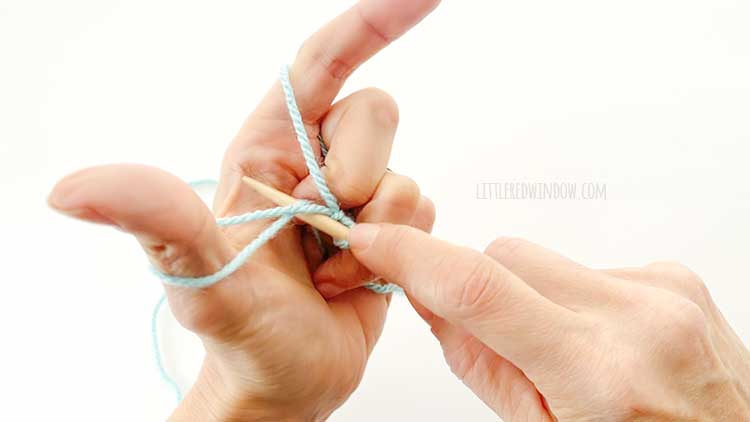 Two hands in front of a white background holding one knitting needle and light blue yarn showing german twisted cast on step 3