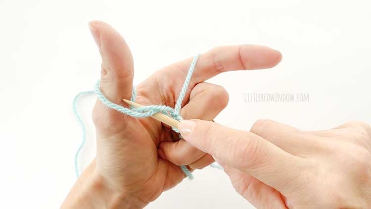 Two hands in front of a white background holding one knitting needle and light blue yarn showing german twisted cast on step 4