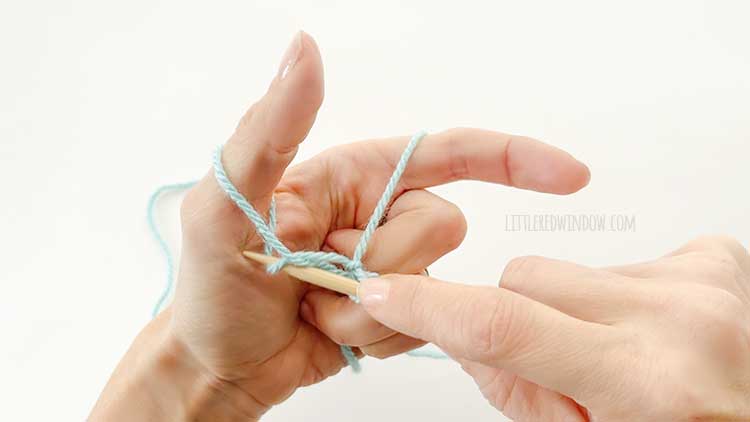Two hands in front of a white background holding one knitting needle and light blue yarn showing german twisted cast on step 5