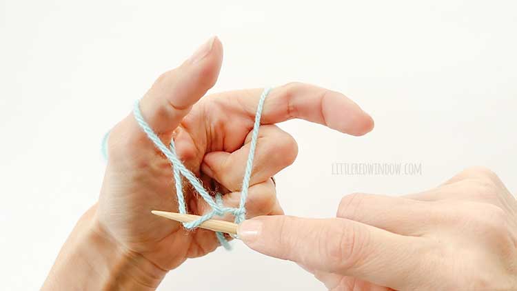 Two hands in front of a white background holding one knitting needle and light blue yarn showing german twisted cast on step 6