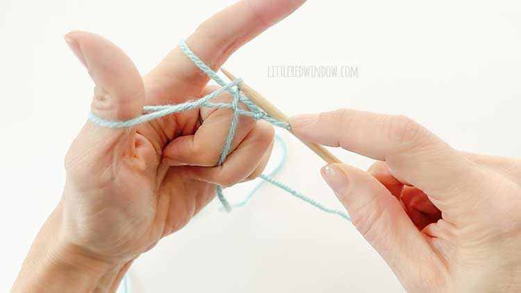 Two hands in front of a white background holding one knitting needle and light blue yarn showing german twisted cast on step 7