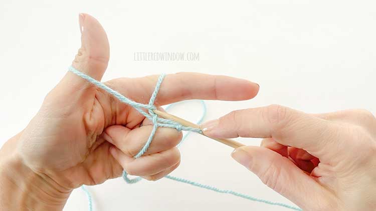 Two hands in front of a white background holding one knitting needle and light blue yarn showing german twisted cast on step 8