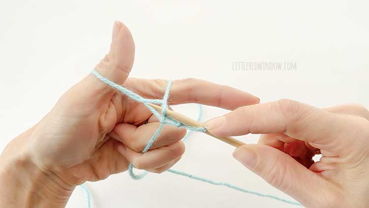 Two hands in front of a white background holding one knitting needle and light blue yarn showing german twisted cast on step 9