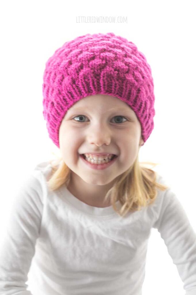 smiling girl looking at the camera and wearing a knit hat that looks like a raspberry