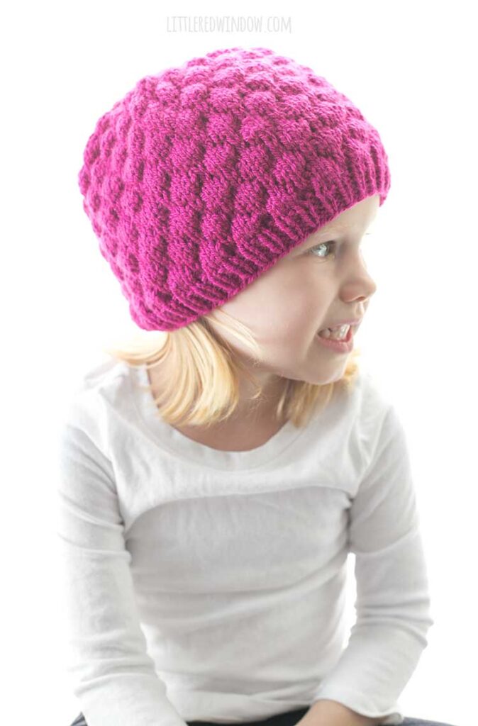 girl in white shirt wearing knit raspberry hat and looking off to the right