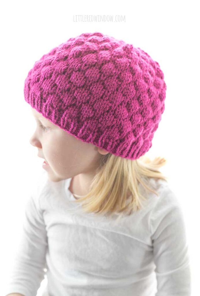 girl in white shirt wearing knit raspberry hat and looking off to the left