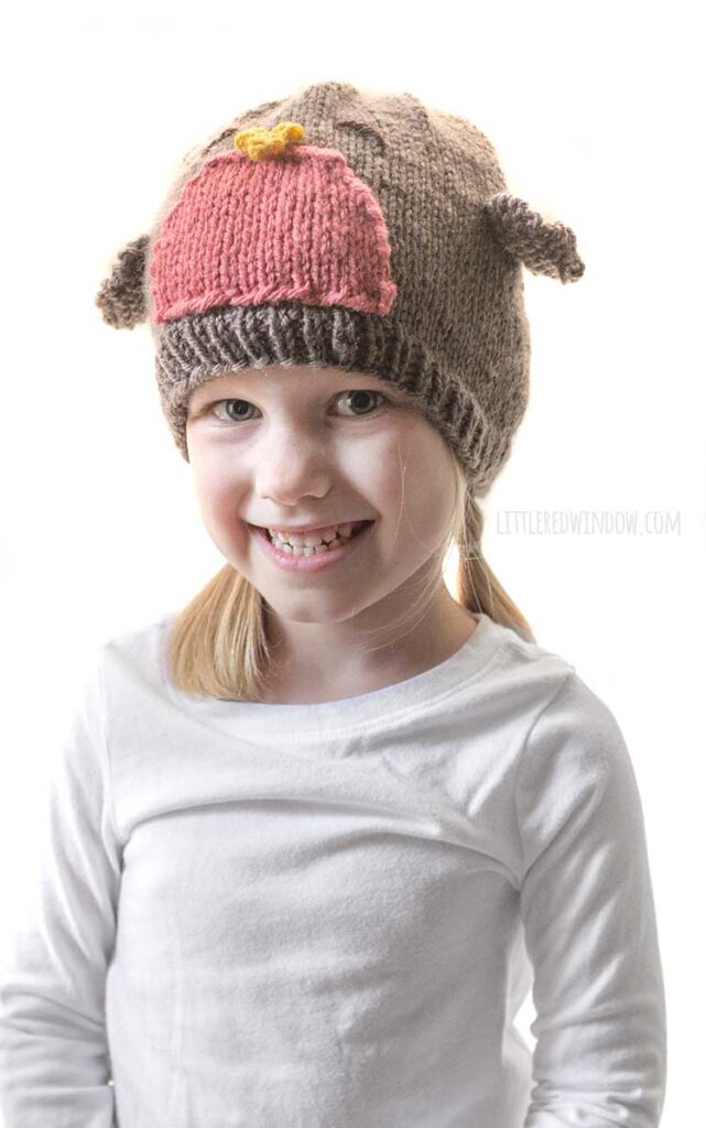 smiling girl in white shirt and blonde braids wearing a brown knit hat that looks like a robin redbreast