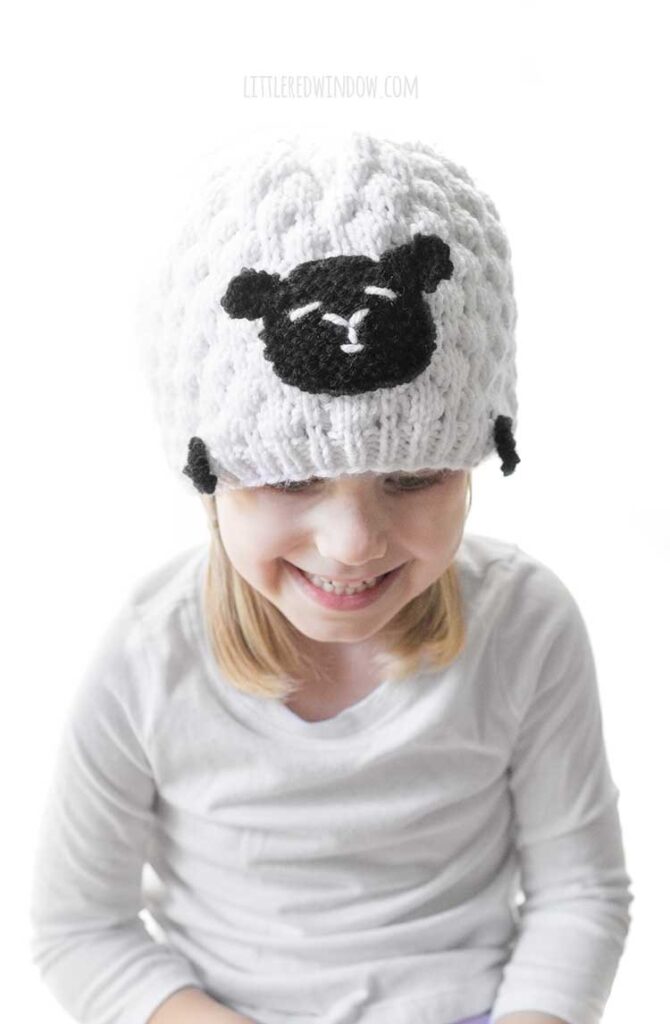 girl in white knit hat the looks like a sheep looking down and smiling