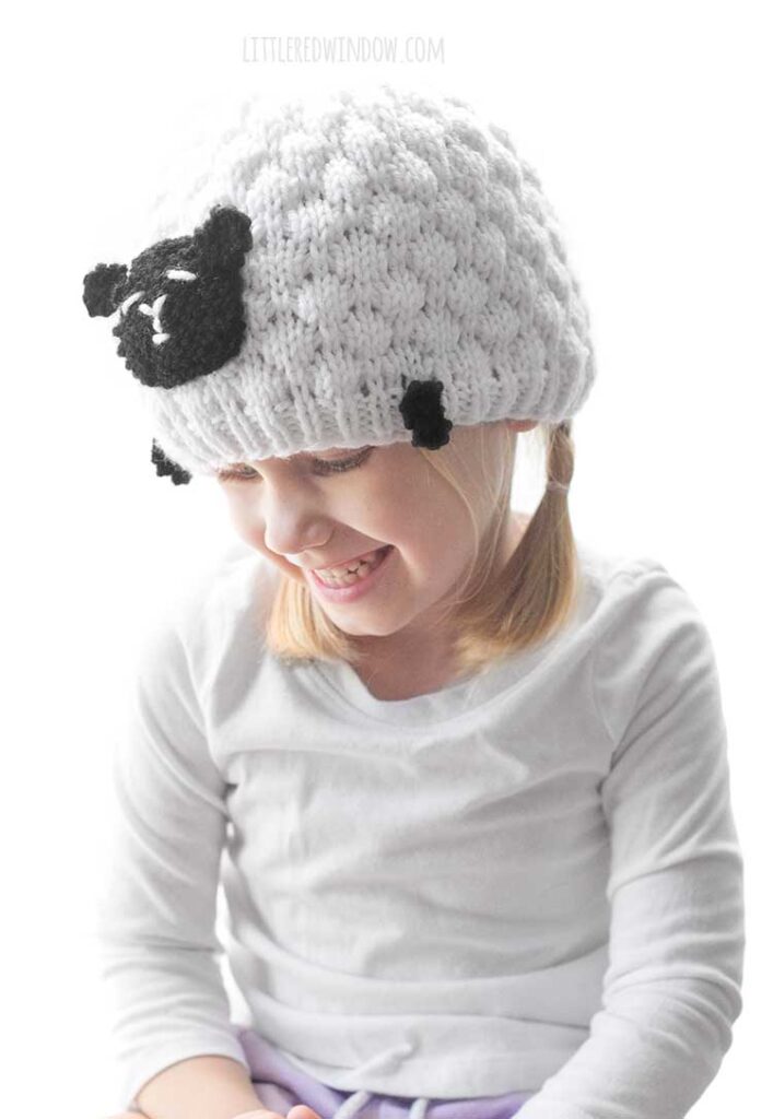 girl with braids wearing a bumpy white knit hat that has black sheep face and feet
