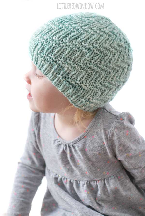 girl looking up and back in a light gray baby shirt and wearing a mint colored knit hat with woven zig zag pattern
