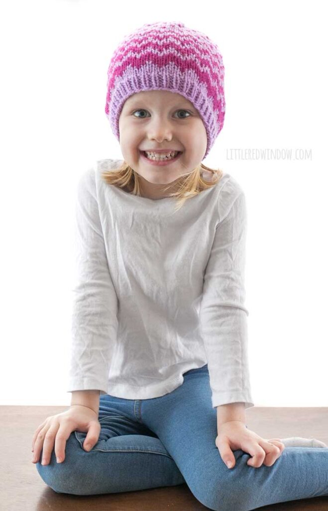 smiling girl in white shirt wearing lavender knit evaporation hat with raspberry colored zig zag pattern
