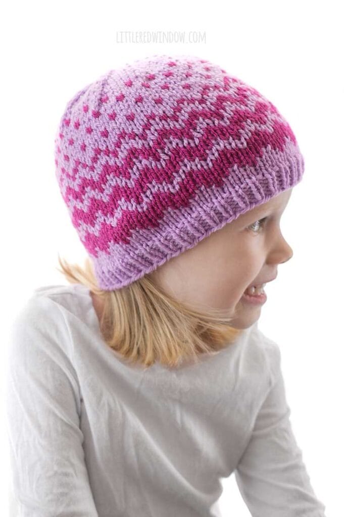 little girl in white shirt looking off to the right and wearing lavender knit hat with decreasing width zig zags in raspberry