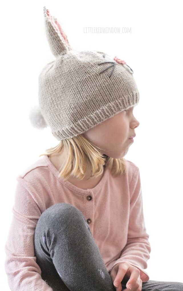 side view of little girl wearing tan and pink bunny hat with white fluffy pom pom tail on the back