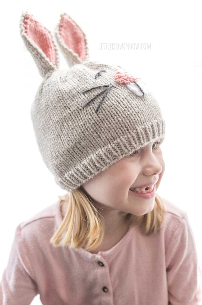 smiling girl wearing tan bunny hat with pink ears and nose looking off to the right