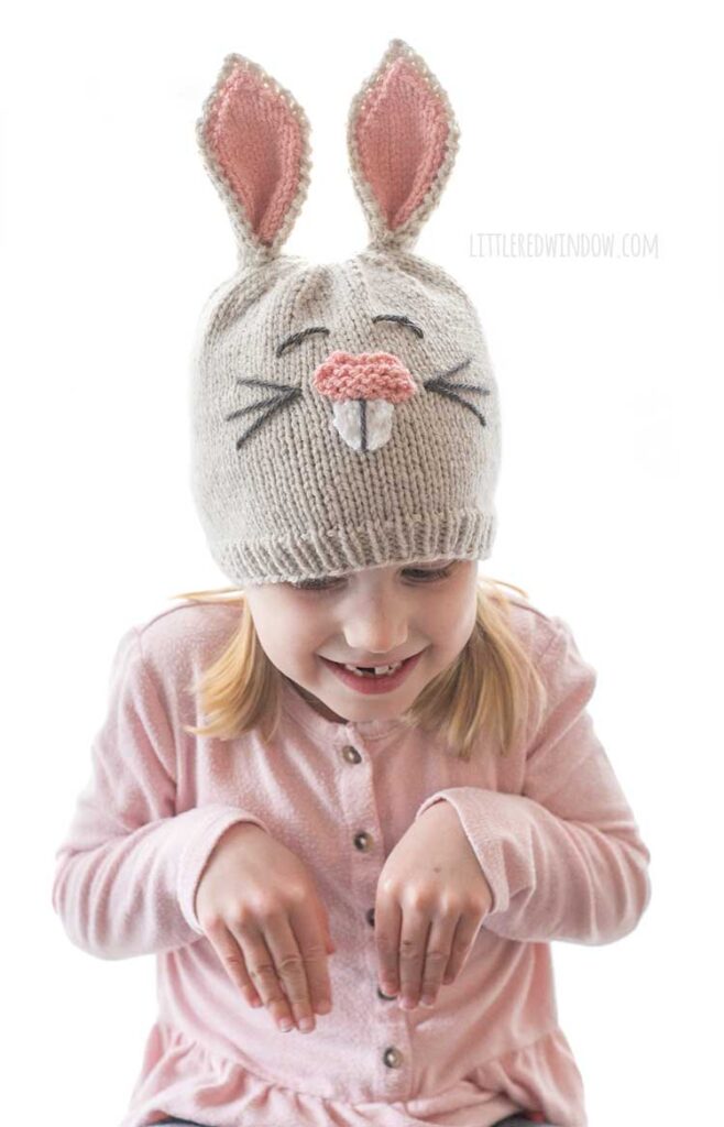 little girl in pink shirt pretending to be a bunny and holding her paws up while wearing a tan knit bunny hat with pink nose and ears