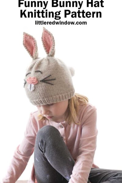 little girl in pink shirt looking down at the ground and wearing a tan knit bunny hat with ears and a tail