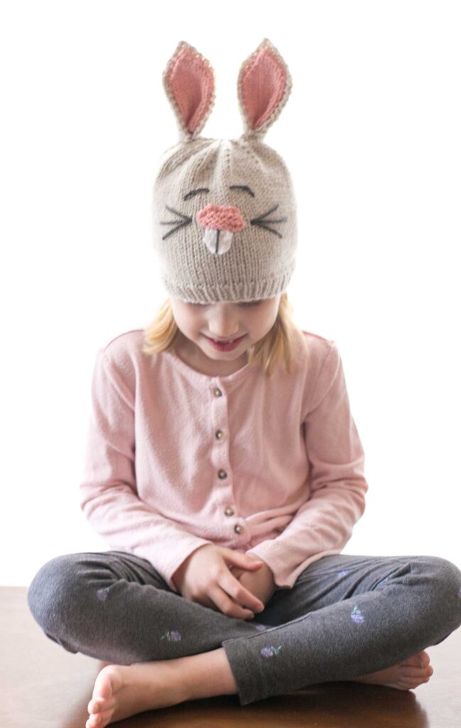 little girl in tan and pink funny bunny hat knitting pattern looking down at her lap