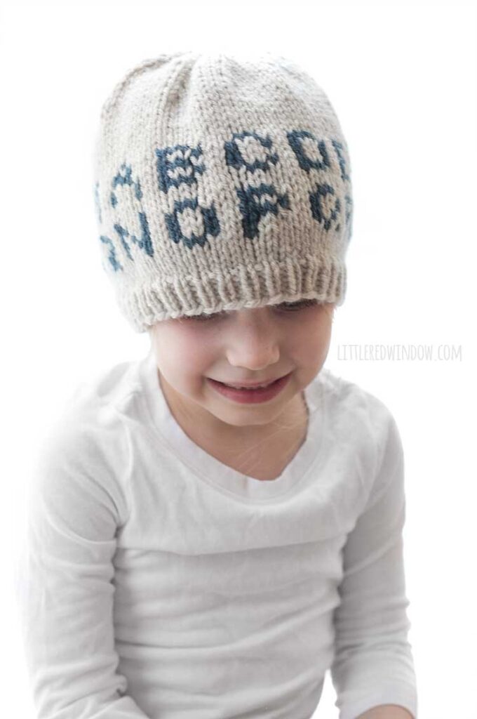 little girl in white shirt wearing tan knit hat with the alphabet on in in dark blue looking down at her lap