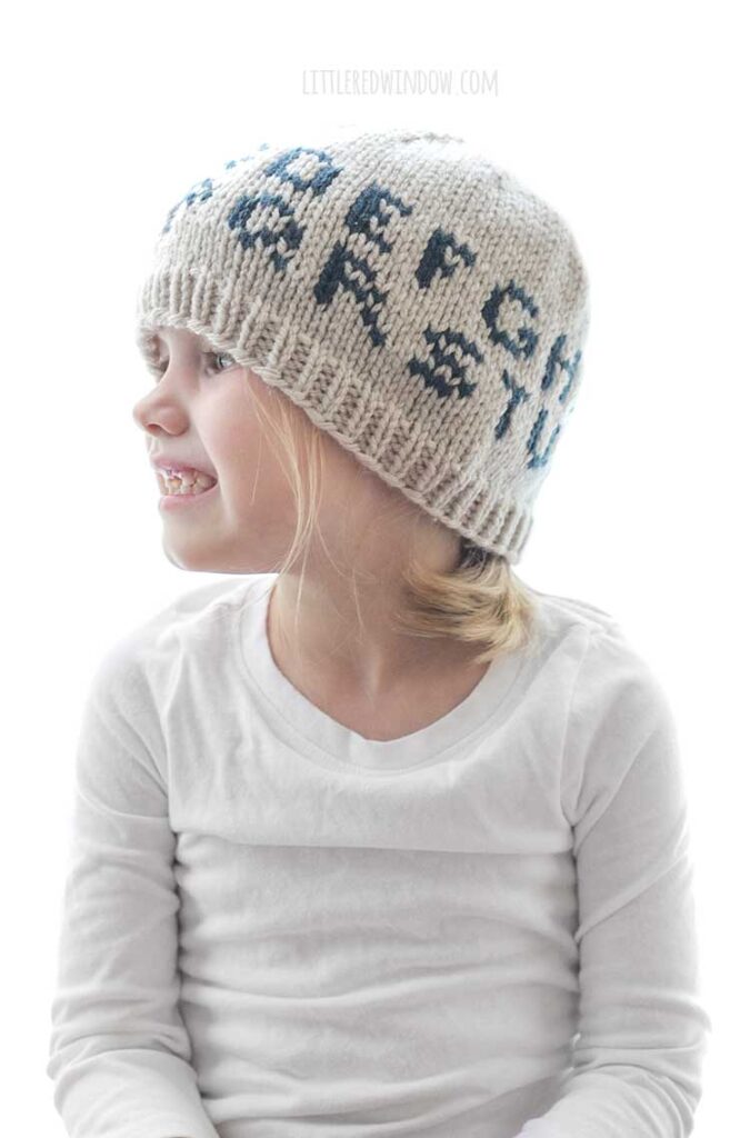 little girl in white shirt wearing tan knit hat with the alphabet on in in dark blue looking off to the left