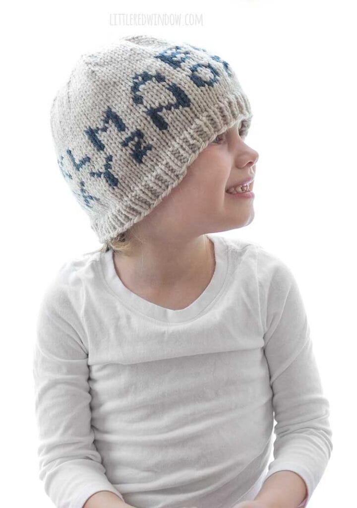 little girl in white shirt wearing tan knit hat with the alphabet on in in dark blue looking off to the right