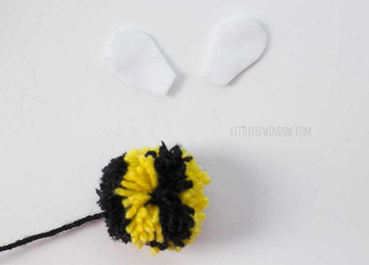 yellow and black striped yarn pom pom next to two white felt bee wings on a white tabletop