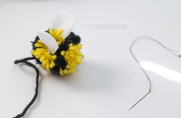 yellow and black striped yarn pom pom with two white felt wings sewn into it to look like a bee with a needle and black thread next to it on a white tabletop