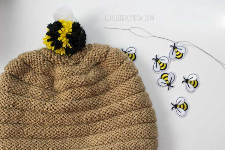 Bee pom pom atttached to the top of a tan knit hat with small bee patches and a needle and thread on the table next to it
