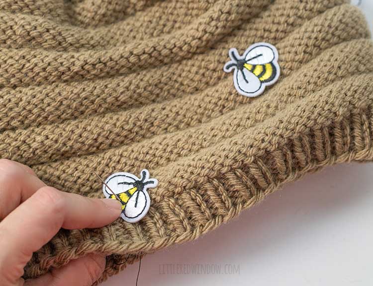 a hand sewing a small bee shaped patch to the brim of the tan knit beehive hat