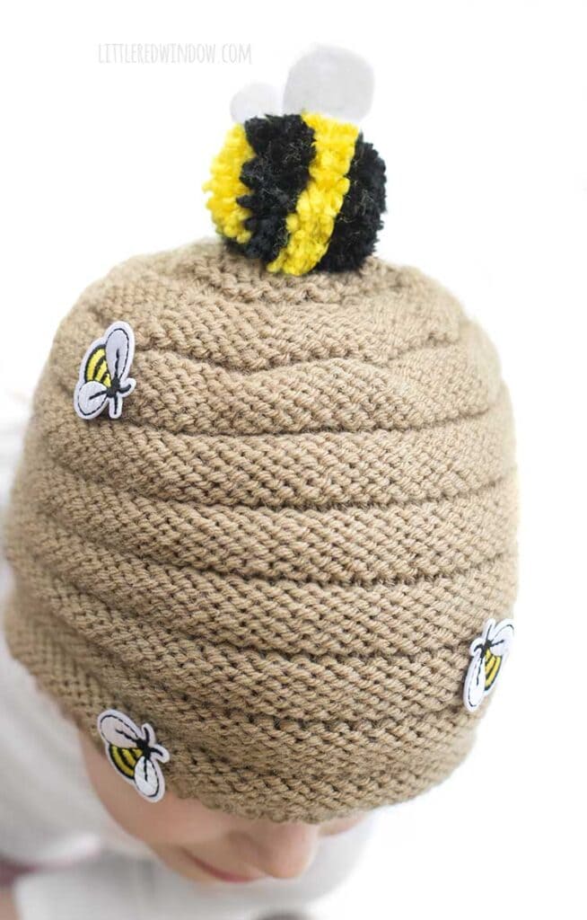 side view of little girl wearing tan knit beehive hat with a black and yellow striped bee pom pom on top and small bee patches sewn on the sides