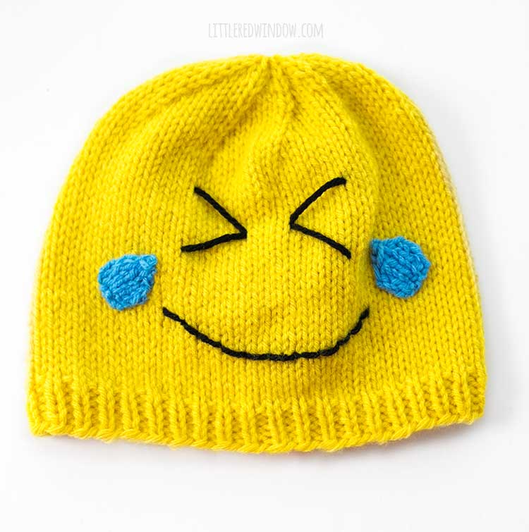 yellow knit emoji hat with squinting eyes two blue teardrops and a smile on a white background