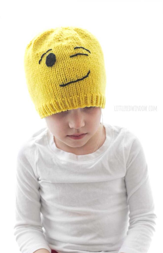 child in white shirt wearing a yellow knit winking emoji hat and looking down at her lap