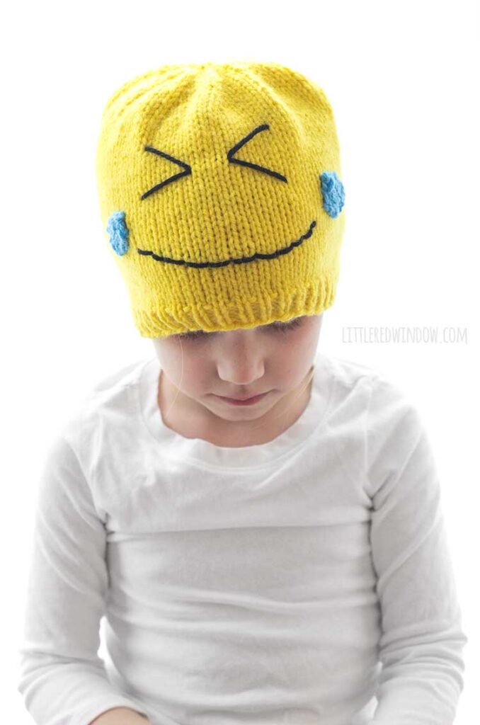 child in white shirt wearing a yellow knit laughing emoji hat and looking down at her lap