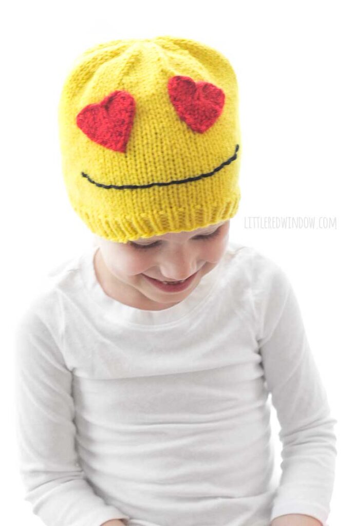 smiling child in white shirt wearing a yellow knit heart eyes emoji hat and looking down at her lap